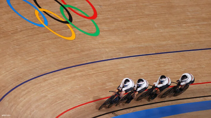 A team of four cyclists, wearing matching white uniforms and aerodynamic helmets, rides closely together in a line on a wooden indoor velodrome track. They are navigating a steeply banked curve marked with red, blue, and black lines. Above them, the rings of the Olympic logo are visible on the wooden surface.