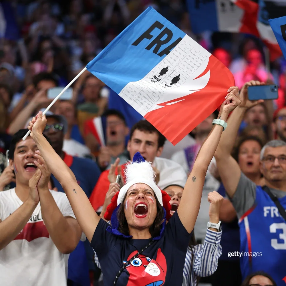 Mujer sosteniendo una bandera de Francia que dice FRA mientras anima. Detrás de ella, un grupo de aficionados franceses está sentado.