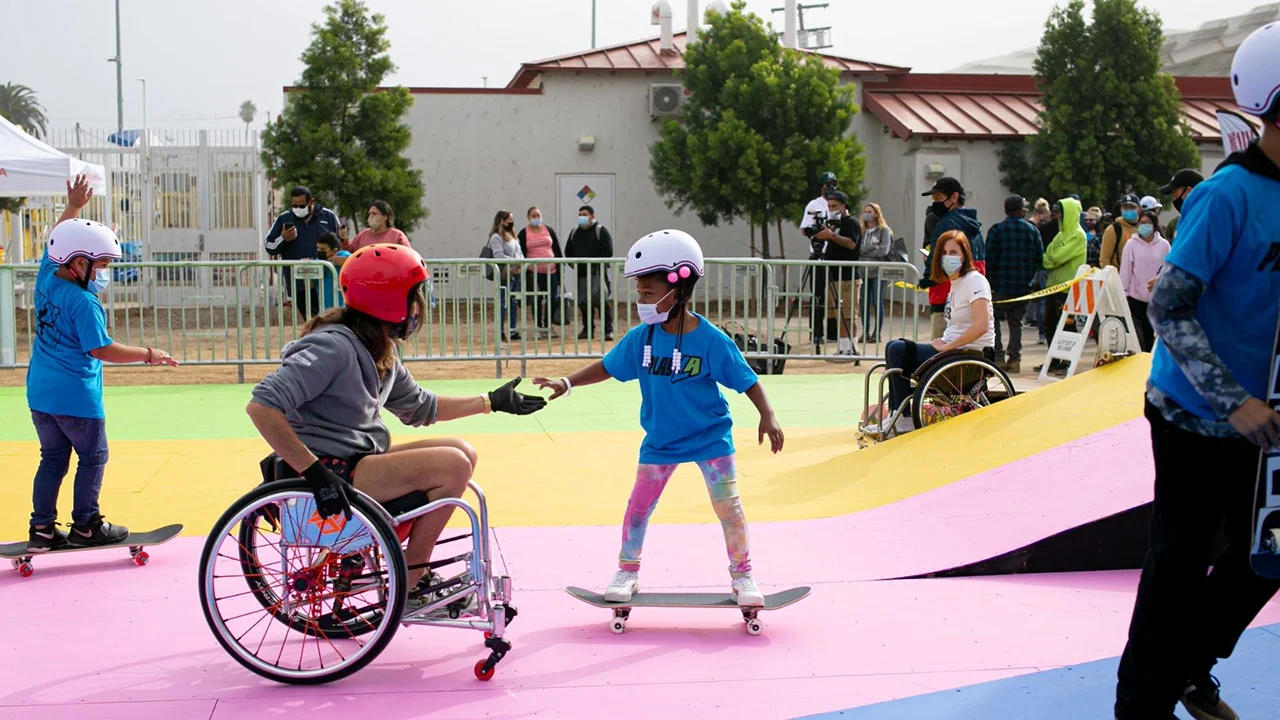 Baloncesto adaptado en silla de ruedas para jóvenes en el evento PlayLA. La imagen captura a una niña montando en skate y extendiendo la mano hacia una mujer en silla de ruedas con casco.