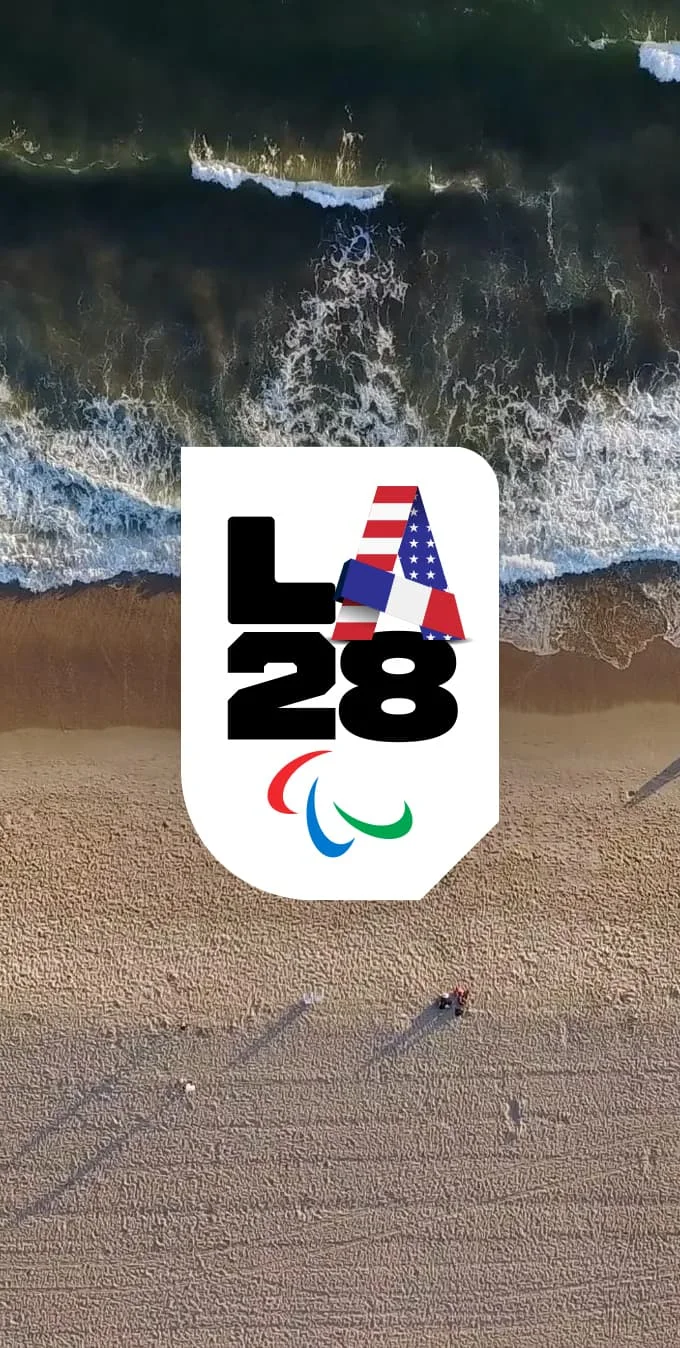 Aerial photograph of a Los Angeles beach with the sand and crashing waves shown from above, and the LA28 Paralympic Handover A Emblem placed on top of the image on a white background. 