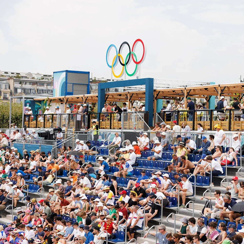 Aficionados en las gradas de los Juegos Olímpicos de París 2024, con una sección de terraza de madera en la parte superior y los anillos olímpicos sobre el techo 