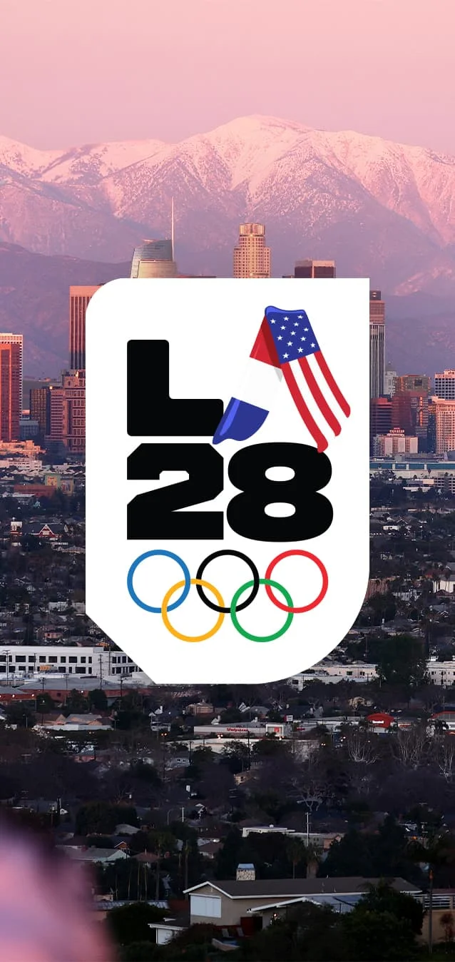 A photograph of the Los Angeles skyline with a sunset and the mountains in the background and the cityscape and a shadow of a person in the foreground. Over the image is the LA28 Handover ‘A’ custom emblem which features the flags of France and the United States overlapped to form an ‘A’. The ‘A’ is next to a static and bold ‘L’ with a number ‘2’ and a number ‘8’ underneath the ‘L’ and the ‘A’ to form the base of the LA28 emblem. The Olympic rings logo is underneath the LA28 ‘Handover A’ custom emblem over a white background.  