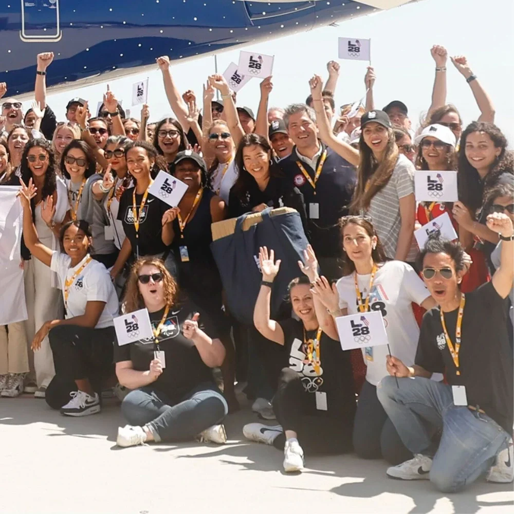 A group of LA28 employees and LA Mayor Karen Bass holding the LA28 dual emblem flag in front of a Delta Olympic Flag Flight plane
