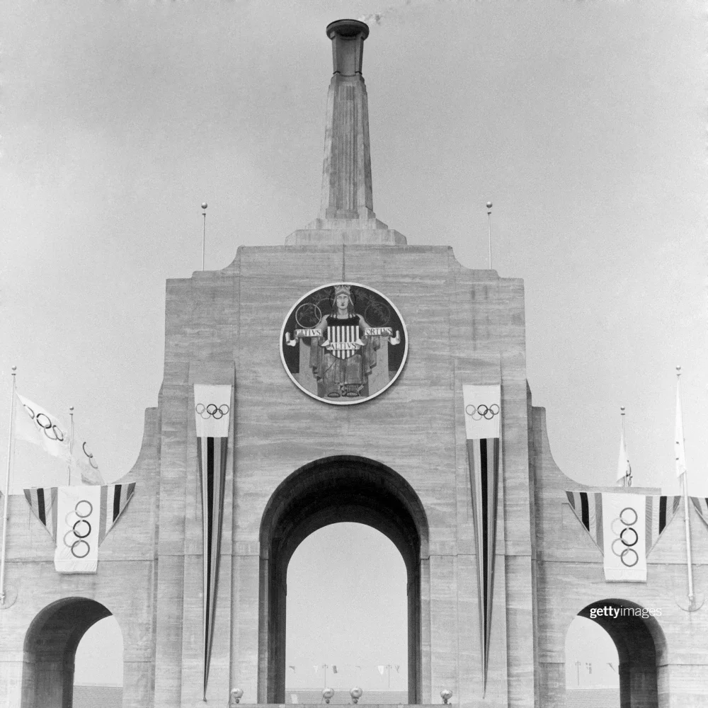Black and white photo of LA Memorial Coliseum displaying a lit Olympic cauldron and eight Olympic flags during the 1932 Games