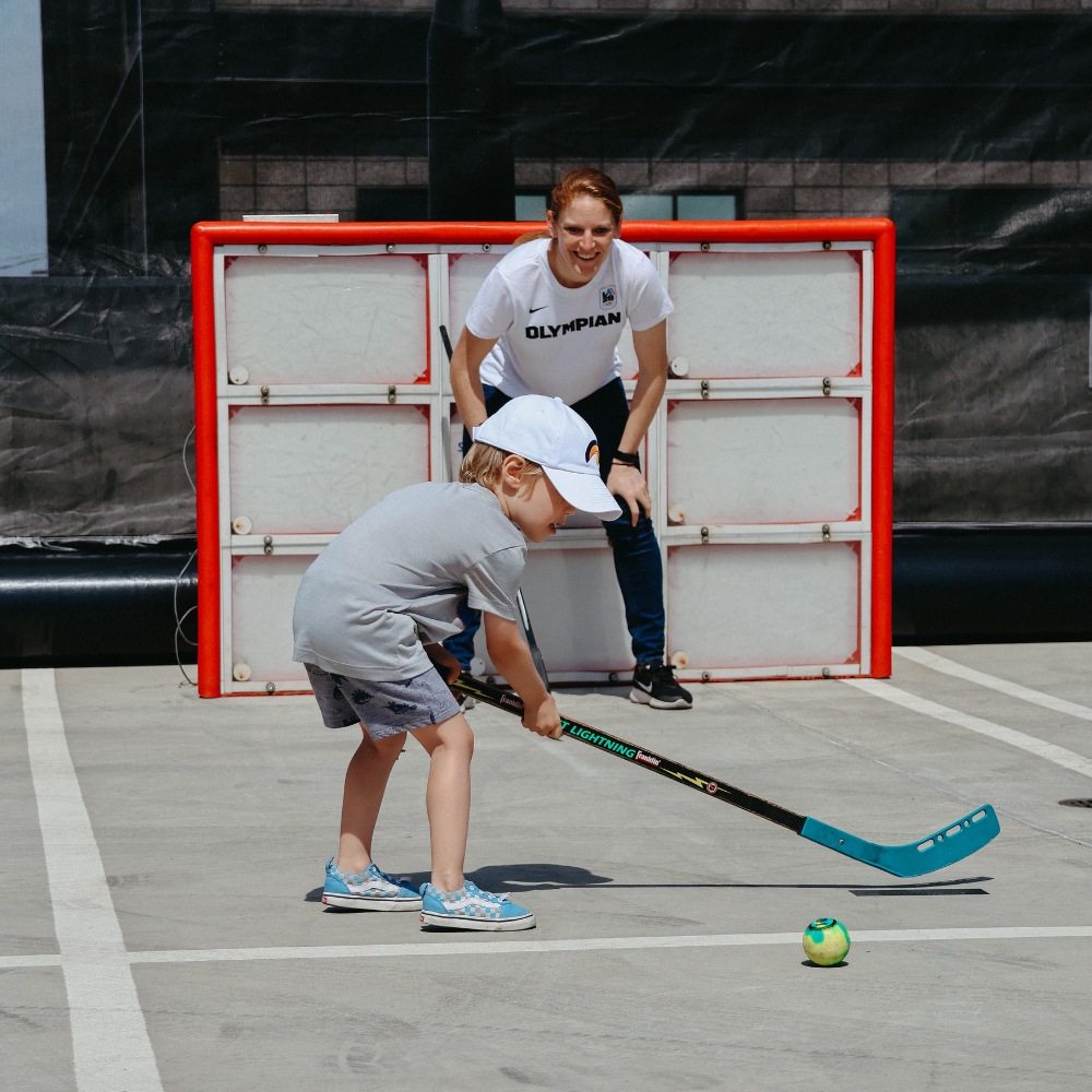 Photo of Winter Olympian, Ice Hockey Goalie Molly Schaus guarding a hockey goal while a young kid holds a hockey stick