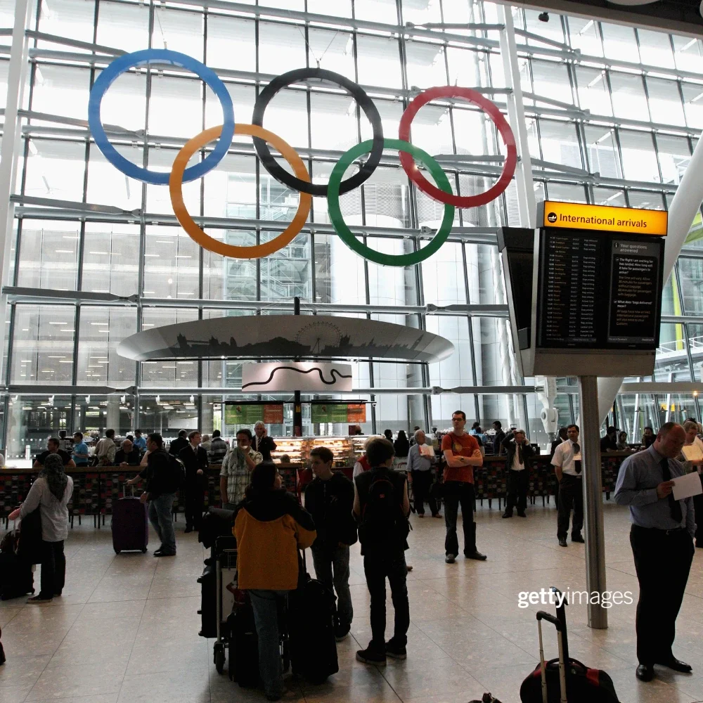 Travelers at an airport with the Olympic rings showcased in front of window panes at the airport. A sign with International arrivals is on the right side of the photograph.
