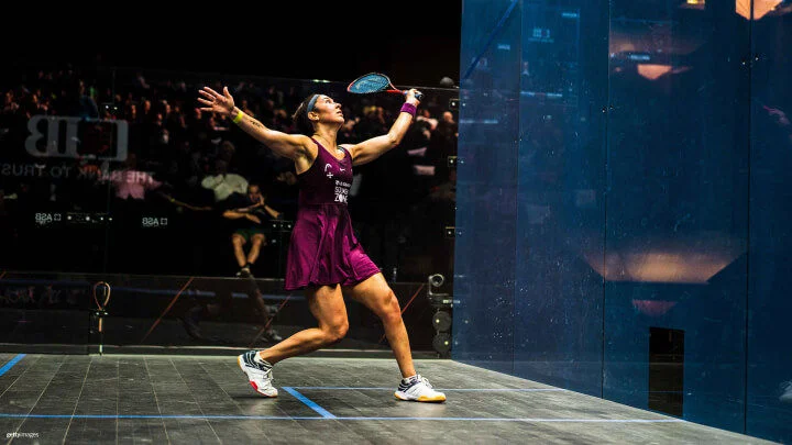 Una jugadora de squash femenina, con un vestido morado, zapatillas blancas, una muñequera y una cinta para la cabeza, se prepara para golpear la pelota con su raqueta. Está en una postura dinámica con su brazo derecho extendido hacia atrás y alto, listo para golpear, mientras que su brazo izquierdo está extendido para mantener el equilibrio. La cancha está rodeada por paredes de cristal, a través de las cuales se puede ver a una multitud de espectadores observando.