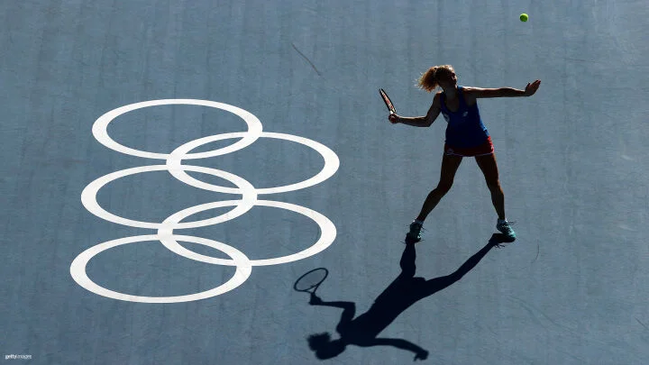 A female tennis player is captured mid-action on a blue court, preparing to hit a tennis ball in the air. The Olympic rings are prominently displayed on the court to her left. The lighting creates a distinct shadow of the player on the court, emphasizing her stance and the raised racket. She is wearing a blue tank top, red shorts, and teal shoes.