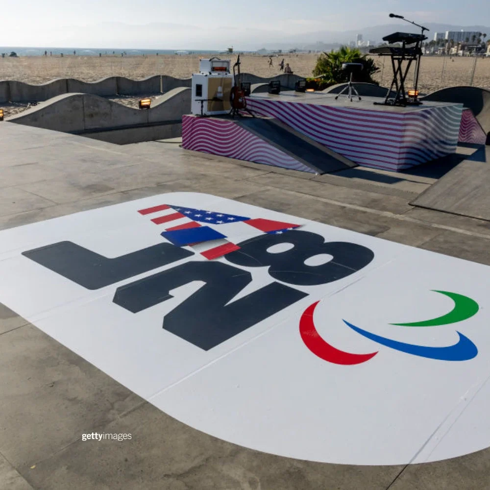 Photo of LA28 Paralympic Handover emblem on a graphic on the floor of the Venice Beach Skate Park with the Paralympic agitos underneath it. An empty keyboard stand and microphones are shown in the background