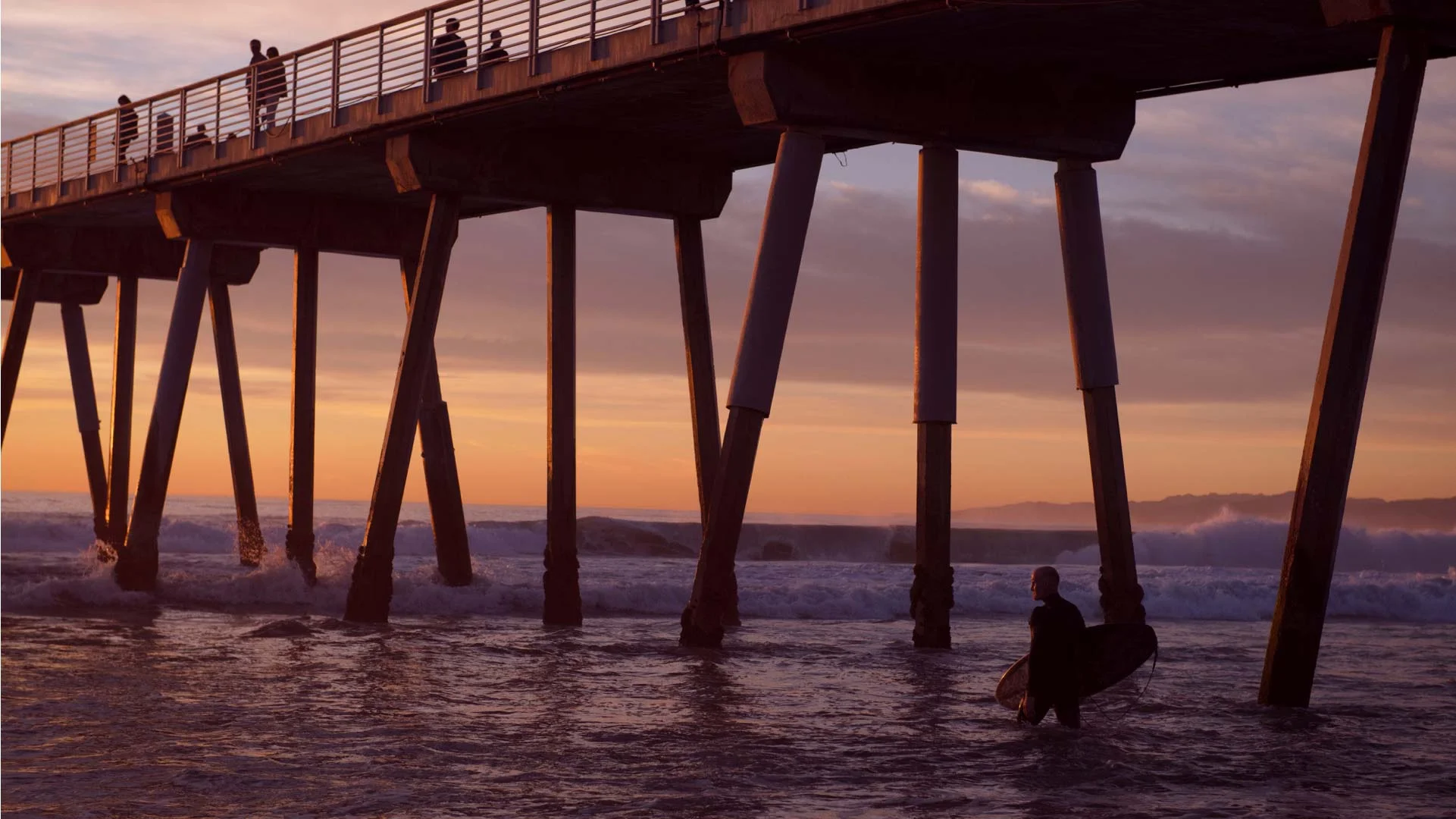 A photo of a surfer in a black wetsuit holding a surfboard under one arm, walking into the ocean, with a pier in the background, the sky is light peach, pink and purple and the text on the photo says Latest Headlines