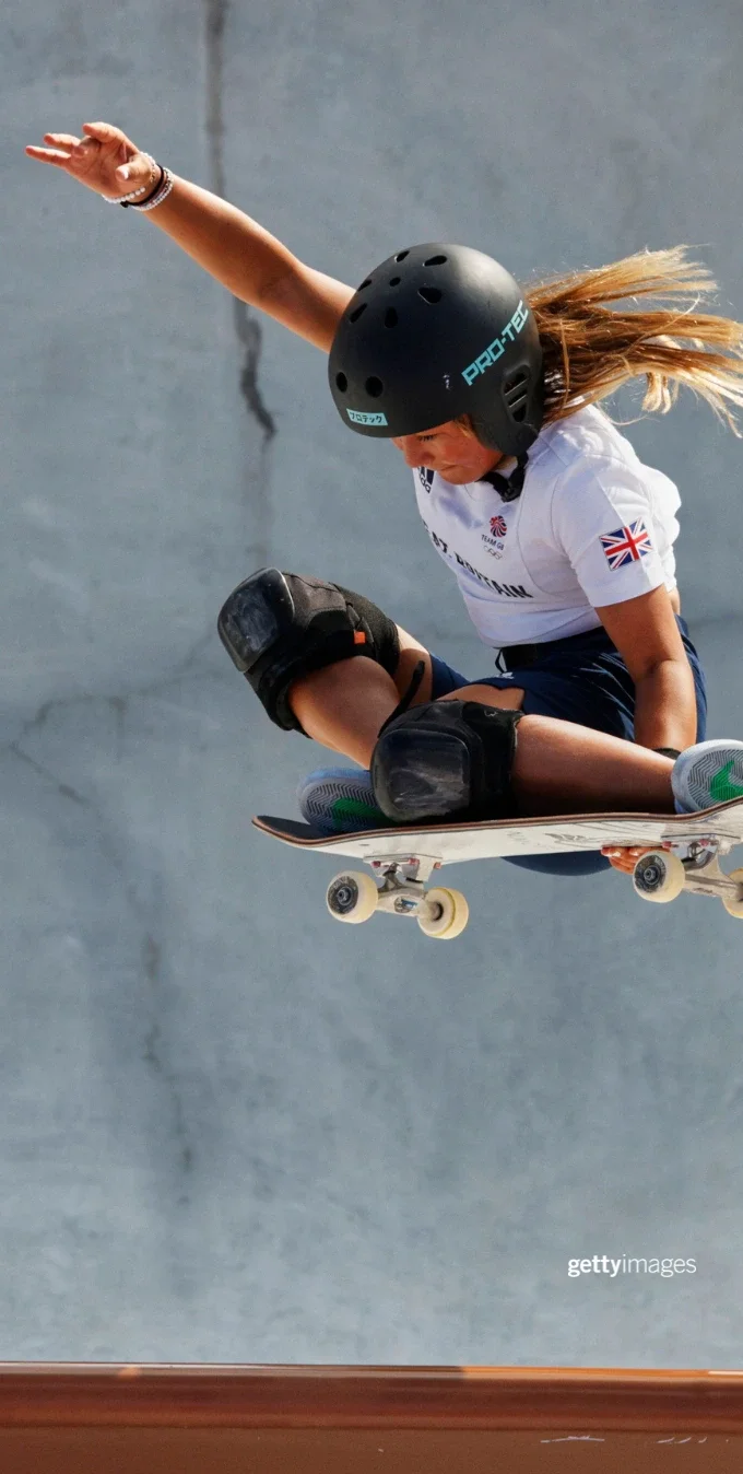 La atleta olímpica Sky Brown en una patineta en el aire mientras usa casco y compite en los Juegos Olímpicos. Al fondo, se ve una pared de concreto gris.