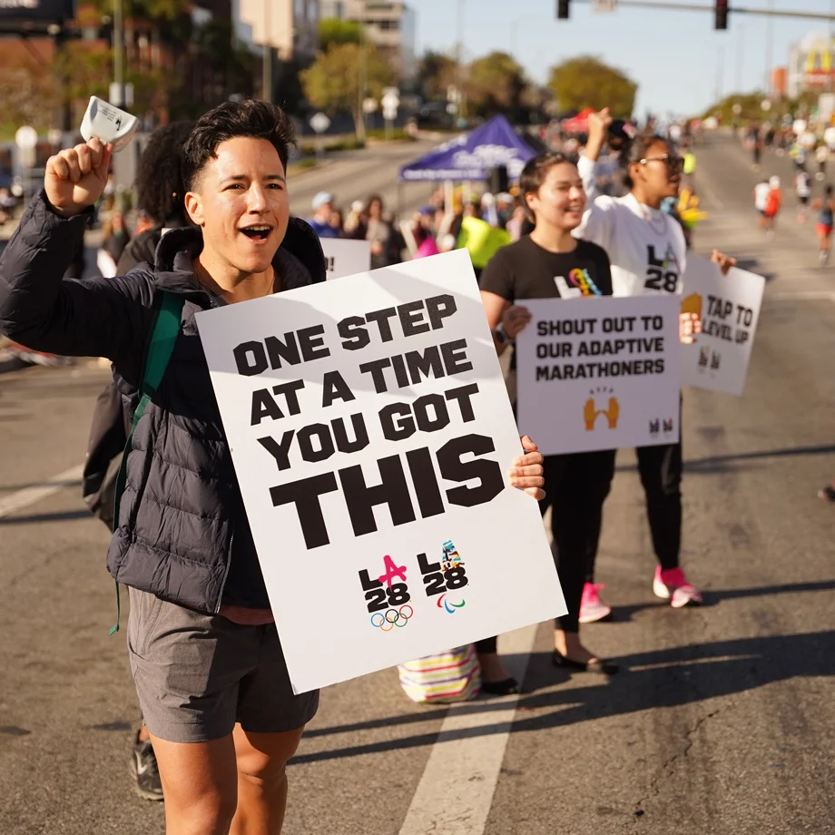 LA28 Employee standing on the side of the marathon route shouting with a sign that says One Step At A Time You Got This