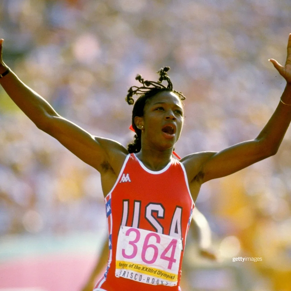 Photo of Team USA Athletics Olympian Valerie Brisco-Hooks celebrating with her hands in the air wearing a red track uniform with the word USA on the front while running at the outdoor track at the LA Memorial Coliseum during the 1984 Games
