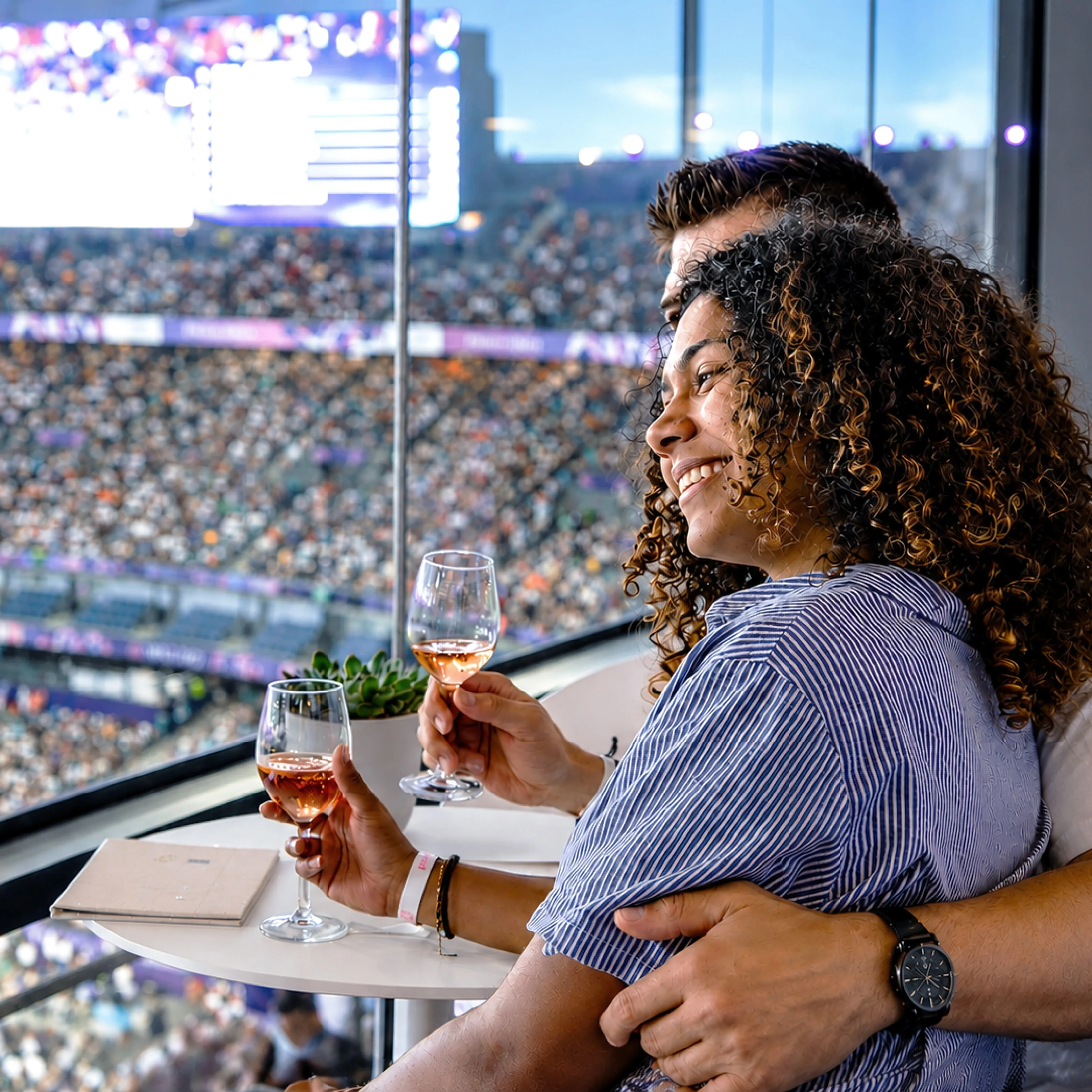 A woman leaning on a man while at the Paris 2024 Games Stade de France Suite while holding glasses of rose wine. 