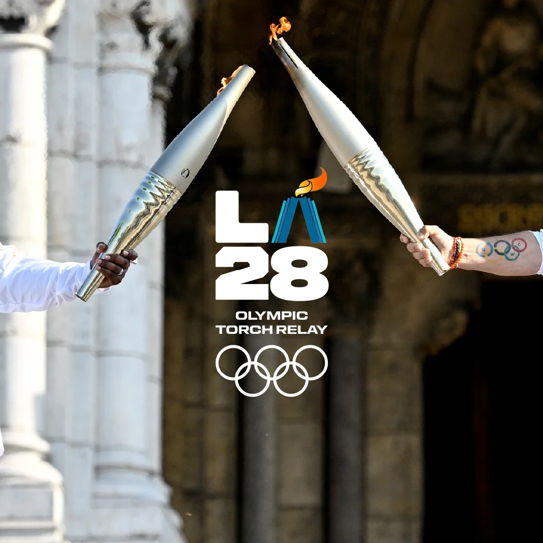 Olympic torch bearers dressed in white long sleeve t-shirts and touching the Olympic torch together in front of columns in the background with the LA28 Olympic Torch Relay emblem superimposed on top