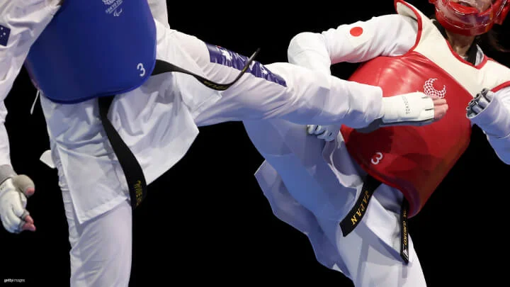 Una imagen cercana de dos atletas Paralímpicos de taekwondo capturados en medio de una pelea durante una competencia, sobre un fondo negro. El atleta de la izquierda lleva un protector de pecho azul y un uniforme blanco con un cinturón negro, ejecutando una patada alta. El atleta de la derecha, con un protector de pecho rojo y un uniforme blanco con la bandera de Japón y un cinturón negro, está bloqueando la patada. Ambos atletas llevan protecciones en la cabeza y guantes.