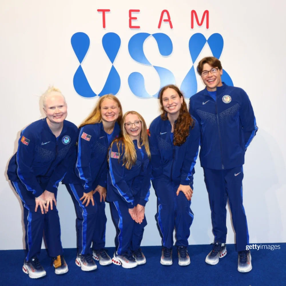 A photograph of paralympian Jack O'Neil and other Team USA paralympians wearing navy blue Team USA uniforms standing in front of a white wall with the blue letters Team USA