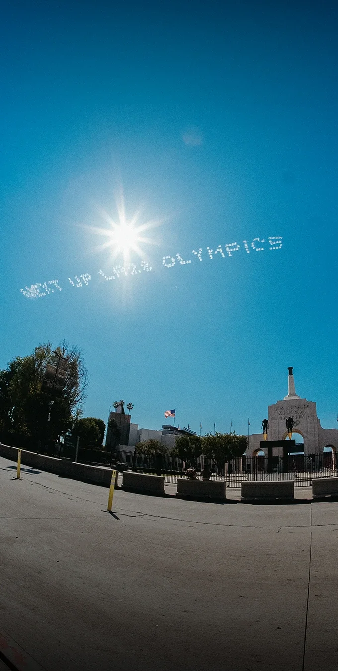 Image of the LA Memorial Coliseum with a blue sky in the background and the words "Next Up LA28 Olympics" in skywriting made by a plane.