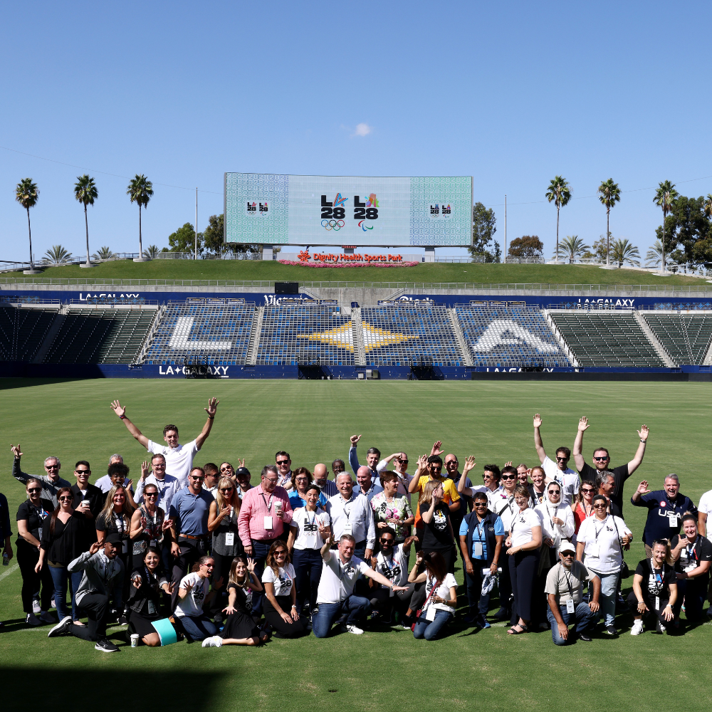 Group photo taken on a soccer field featuring about 50 people smiling and raising their hands