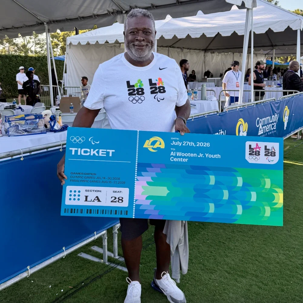 Man wearing a LA28 white t shirt holding a giant ticket in front of a white canopy tent standing on a grass field