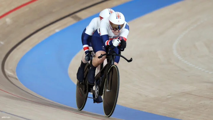 Dos ciclistas están pedaleando juntos en una bicicleta tándem en un velódromo bajo techo. Ambos atletas están vestidos con trajes de ciclismo blancos, azules y rojos del equipo de Gran Bretaña, con cascos aerodinámicos blancos y gafas. El ciclista en la parte delantera sujeta firmemente el manillar mientras que el ciclista de atrás está parcialmente fuera de la vista. Están inclinados hacia adelante en posición aerodinámica mientras atraviesan velozmente la curva de la pista, la cual tiene una superficie de madera clara con marcaciones azules y rojas. El fondo muestra las suaves curvas inclinadas del velódromo. 