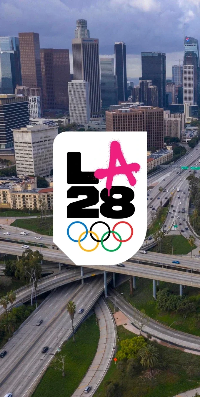 Aerial photograph of downtown Los Angeles and the freeways in the background with the LA28 Street Art Olympic A emblem overlayed on top. 