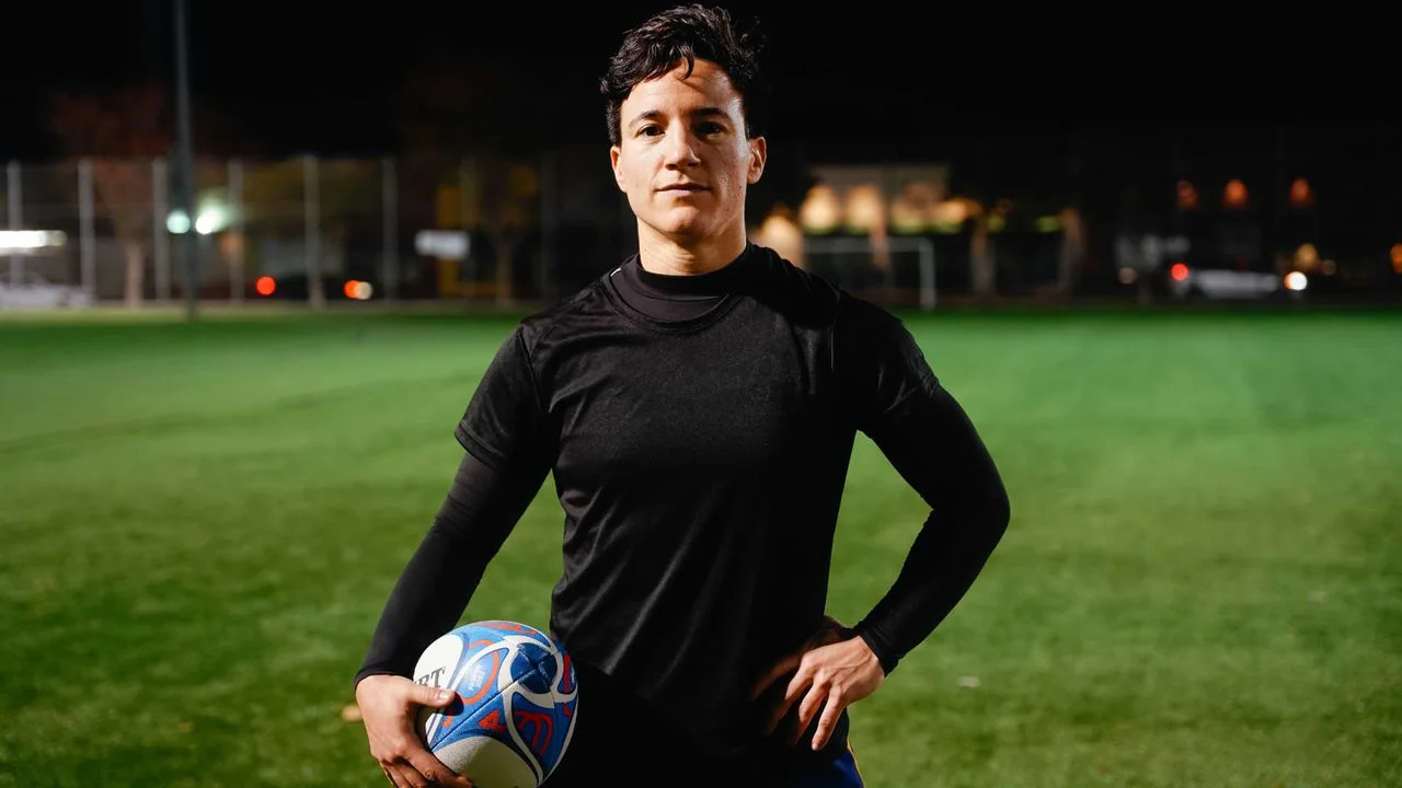 Izzy Cerullo in a black t-shirt with one hand on her hip and one hand on a rugby ball