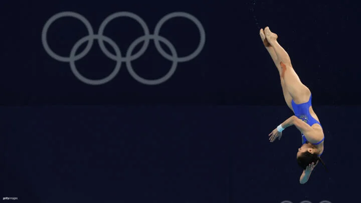 A female diver is captured mid-dive against a dark blue background with the Olympic rings visible in the upper part of the image. She is wearing a blue swimsuit and is upside down, with her body fully extended, pointed toes and her arms positioned by her head.