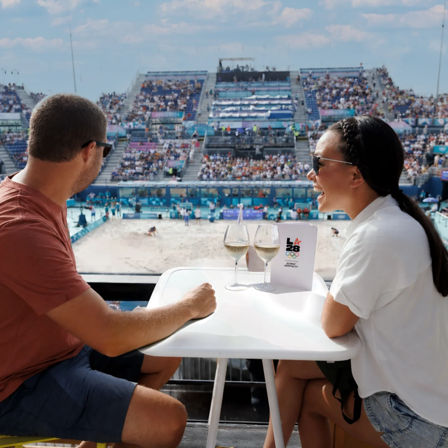 A man and a woman sitting at a table overlooking a beach volleyball court with a LA28 pamphelet standing up on the table. Wine glasses with white wine are on the table. 