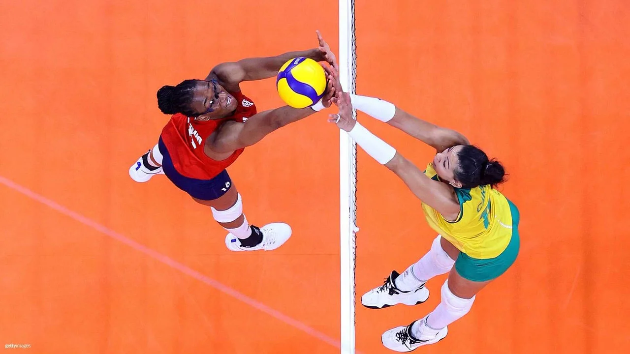 Una jugadora de voleibol del equipo estadounidense, con camiseta roja, intenta rematar la pelota por encima de la red mientras una jugadora del equipo de Brasil, con uniforme verde y amarillo y mangas blancas, salta para bloquearla. Ambas jugadoras están concentradas en el balón, de color amarillo y azul y está marcada con "Tokio 2020". La red tiene el símbolo de los anillos olímpicos y "Tokio 2020" escrito en ella.