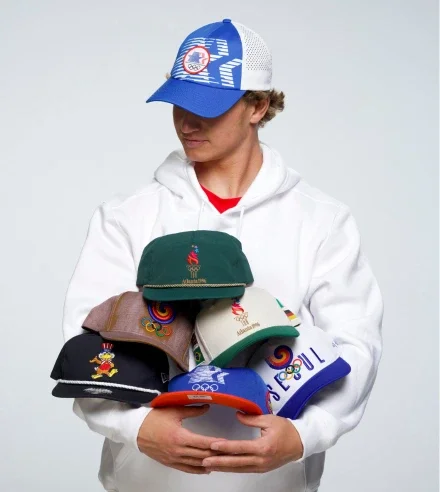 Blonde haired man in an Olympic Heritage Collection baseball hat while wearing a hoodie and holding multiple  Olympic Heritage Collection baseball hats