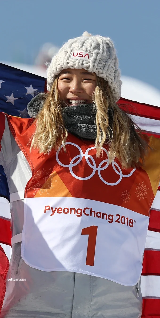 Chloe Kim sosteniendo una bandera americana estirada a la espalda mientras sonríe a la cámara con ropa de snowboard.