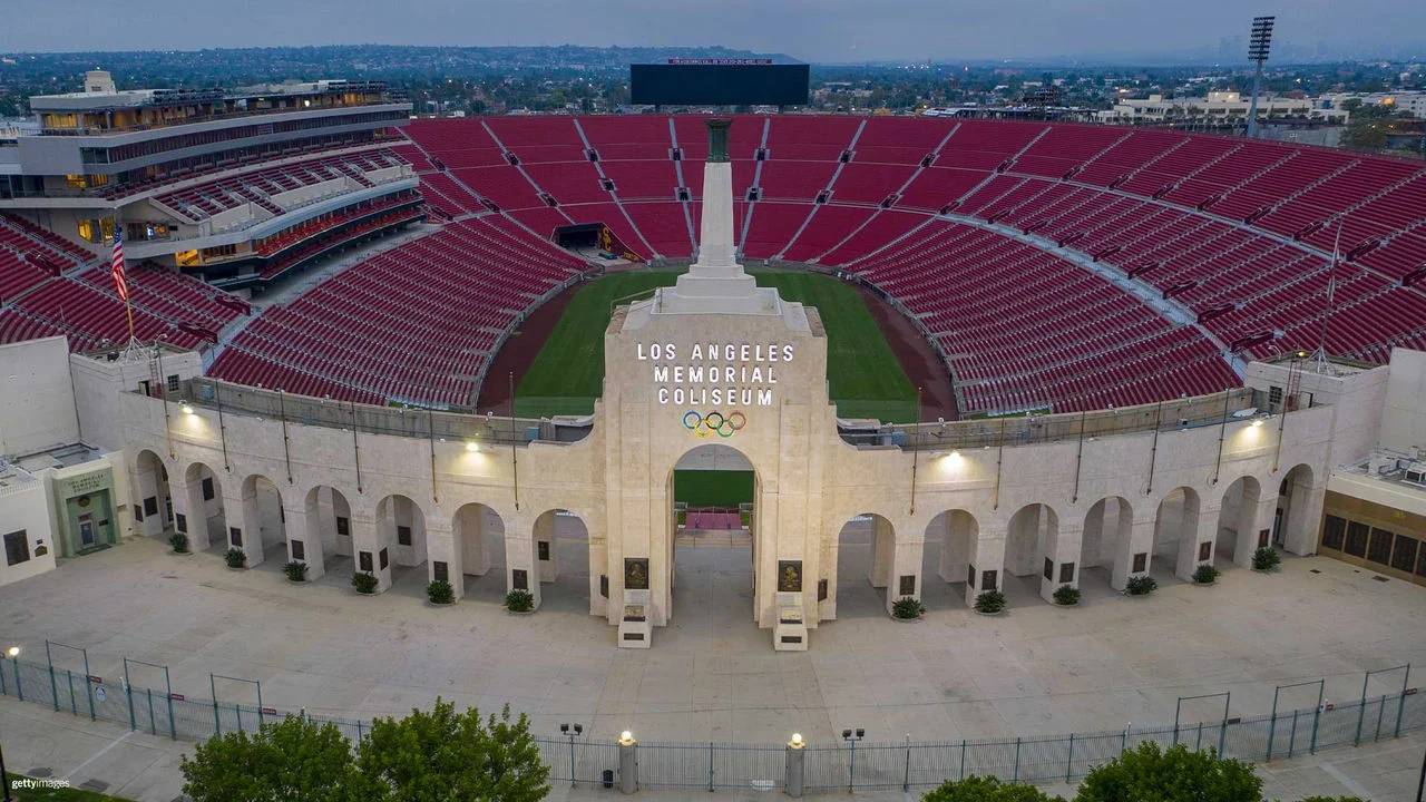 Photograph of the LA memorial Coliseum from the front at dusk with the red seats visible in the background as well as part of the green field and the front gates are tan colored and have lights lit above the stone arches