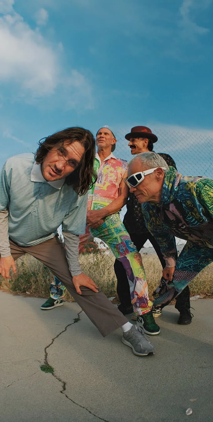 A photograph of the Red Hot Chili Peppers grouped in front of a fence with a blue sky above.