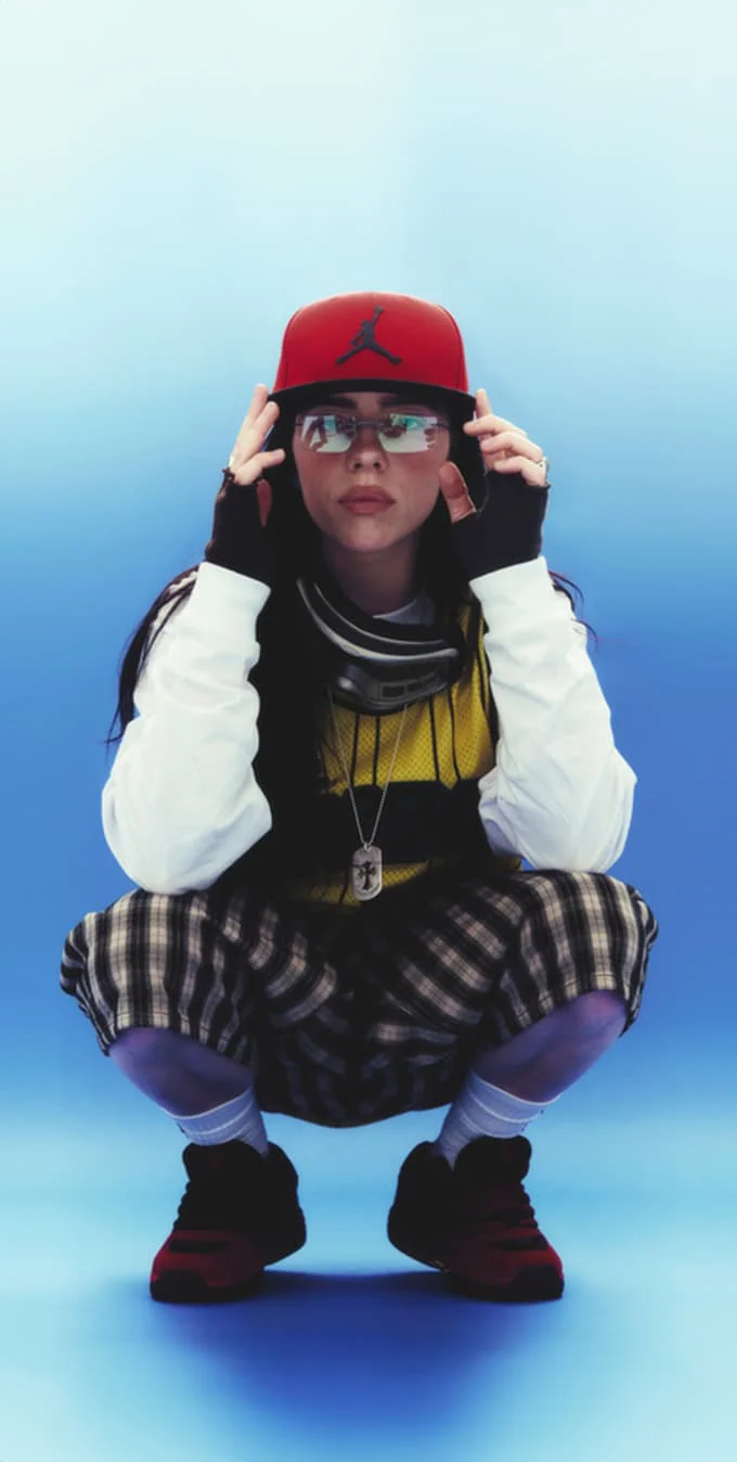 Billie Eilish con una gorra de béisbol roja y gafas de sol agachada delante de un fondo azul.