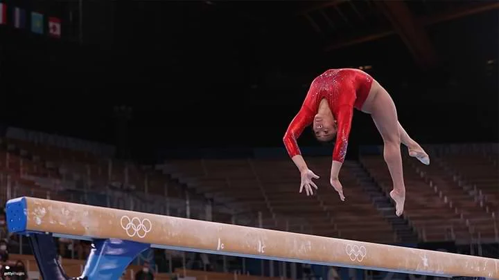 photograph of a female gymnast in a red leotard doing a back flip on a beam caught in mid air