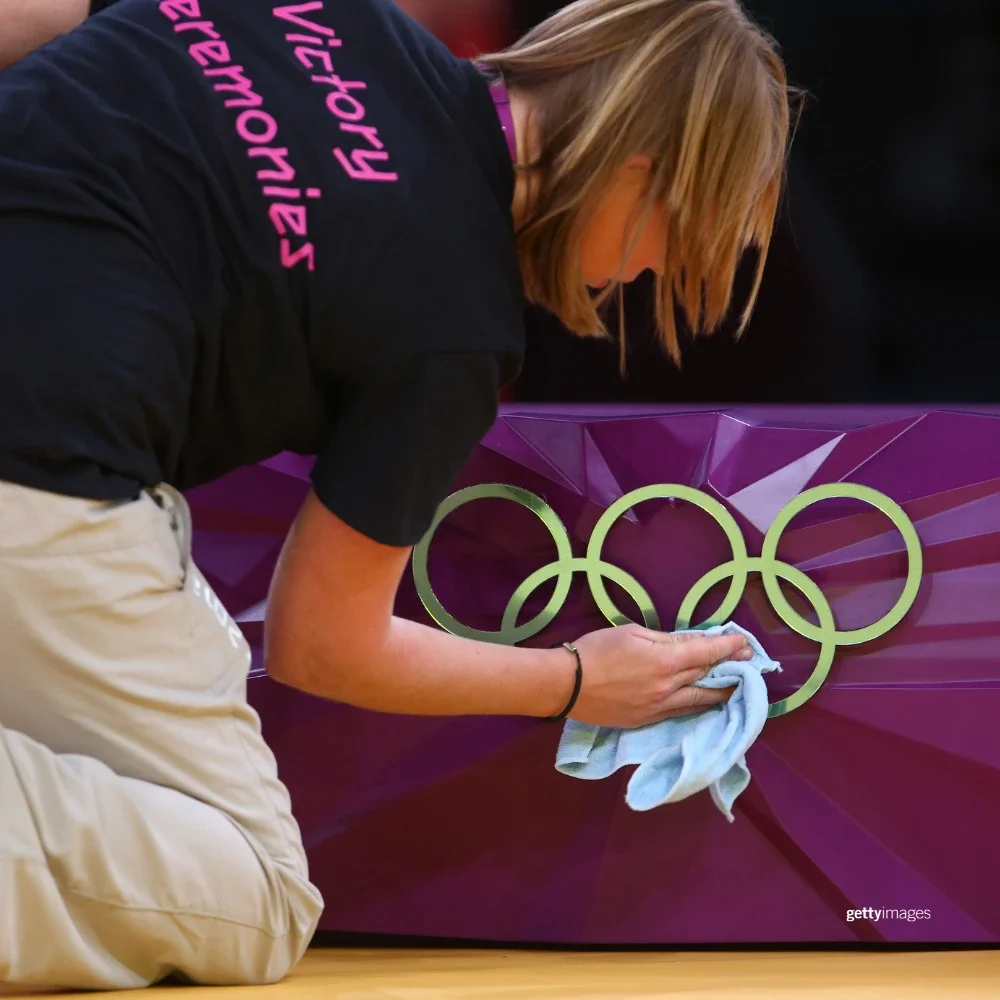 A photograph of a woman kneeling to clean golden Olympic rings on a purple background