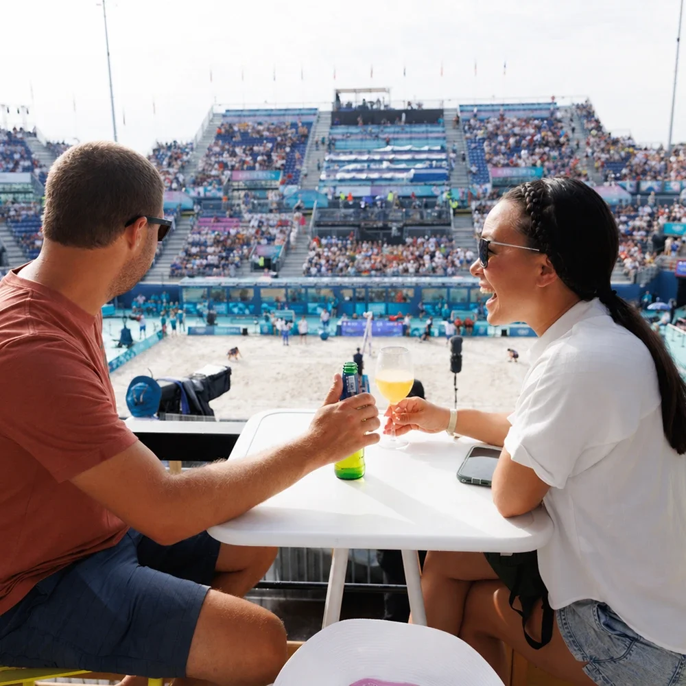 A man with an orange shirt and shorts sitting next to a woman with dark hair and a braid while sitting at a table overlooking a game at an Olympic beach volleyball stadium 