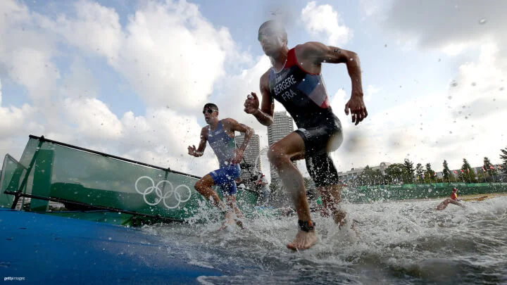 Two triathletes are running out of the water during a competition, with splashes of water surrounding them. The athlete on the right is wearing swim goggles and a black and red triathlon suit labeled "BERGERE FRA," indicating he is from France. The athlete on the left is wearing a blue suit and swim goggles. Behind them, the Olympic rings are displayed on a green banner. The background shows a cloudy sky and some buildings.