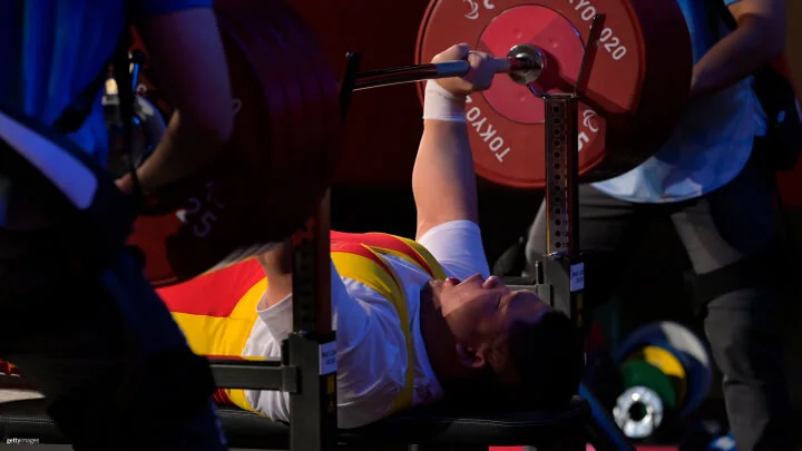 A powerlifter is performing a bench press during a competition. The athlete is lying on a bench, with his mouth open, wearing a yellow and red singlet and a white undershirt, and gripping a barbell loaded with large red weight plates. The scene is dimly lit, highlighting the intensity and focus on the lifter's face. Two spotters are standing on either side, ensuring the athlete's safety.