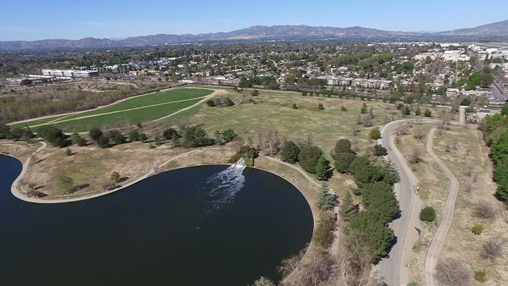 An aerial photograph of the Sepulveda Basin River Recreation Zone, showing a paved road in the forefront, with grass, trees and a small lake in the photograph. Buildings and mountains can be seen in the background of the image depicting the San Fernando Valley.