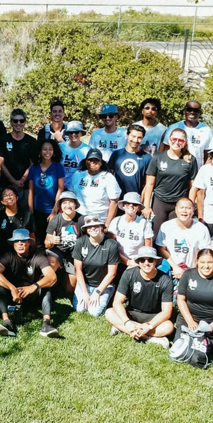 A group photo of many LA28 staff sitting on the grass smiling for a group photo in front of the LA river