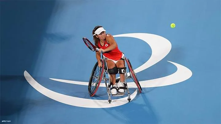 Imagen de una jugadora de tenis Paralímpica femenina sentada en una silla de ruedas, vistiendo una camiseta sin mangas roja y una visera blanca, golpeando una pelota de tenis en una cancha azul con un símbolo de agitos blanco en el suelo