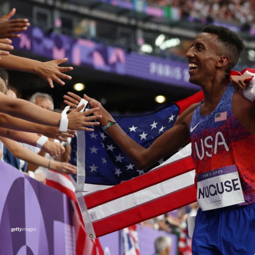 A photo of Yared Nuguse greeting fans in the stand while draped in an American flag after earning his bronze medal at the Paris 2024 Games