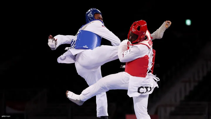 Two athletes in protective gear are engaged in a taekwondo match. The athlete on the left, wearing blue protective gear, is executing a high kick aimed at the athlete on the right, who is wearing red protective gear. Both athletes are mid-air.