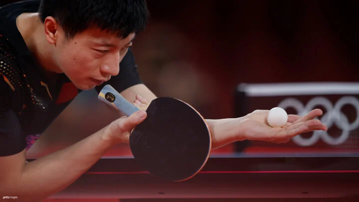 A close-up of a male table tennis player in a black shirt, intensely focused as he prepares to serve. He holds a paddle in his right hand and a ball in his left hand, which is extended outward. The background is blurred, but the Olympic rings can be seen on the net.