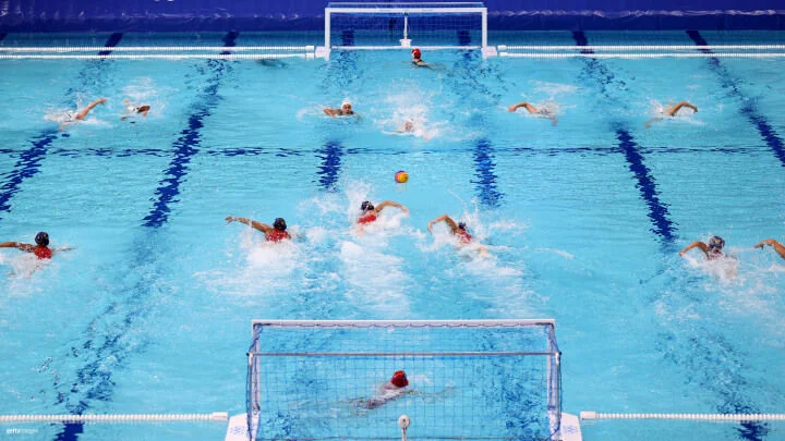 Se lleva a cabo un encuentro de waterpolo en una gran piscina de un color azul brillante. Se ven dos equipos nadando vigorosamente hacia el balón que está flotando en el centro de la piscina. Los jugadores llevan gorros de diferentes colores para distinguir los equipos, con los porteros posicionados en cada extremo de la piscina, protegiendo cada uno una portería. La acción es intensa, con los jugadores generando salpicaduras mientras nadan y se esfuerzan por obtener el control del balón.