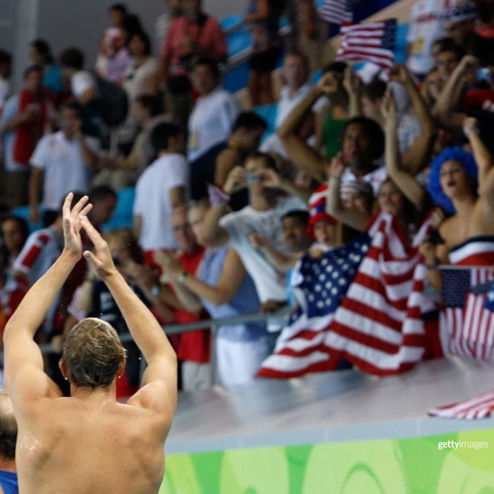 A photograph of a male swimmer from behind with their hands in the air clapping and fans in the background clapping and cheering