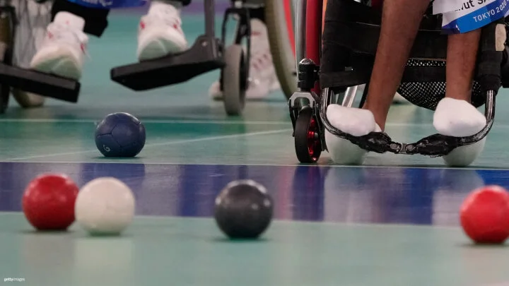 A close-up shot of a boccia game, focusing on the lower part of two athletes in wheelchairs. The athlete on the right is in a red wheelchair with their feet in footrests, wearing white socks. The athlete on the left is partially visible in the blurred background, with white sneakers resting on the footrest. Several boccia balls, including red, blue, white, and black, are scattered on the court.