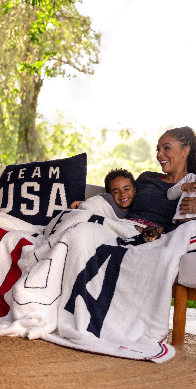 Fotografía de Sydney Leroux, medallista de oro olímpica estadounidense y delantera del equipo Angel City FC, sonriendo al aire libre mientras sostiene un balón y luce un cozie con el diseño de la bandera estadounidense de Barefoot Dreams